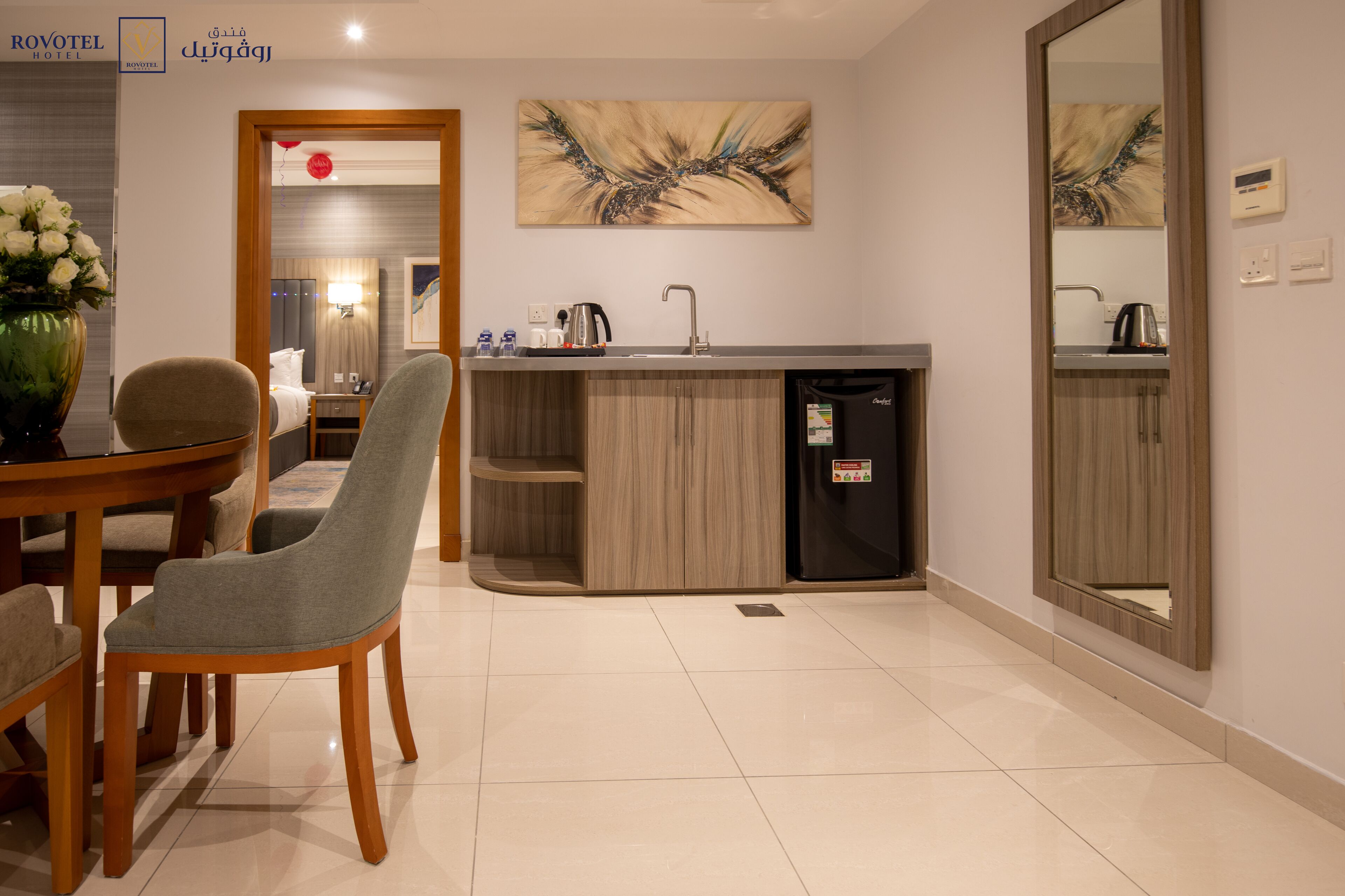 A modern hotel room with a dining area, a kitchen with a sink and a coffee machine, and a large mirror.
