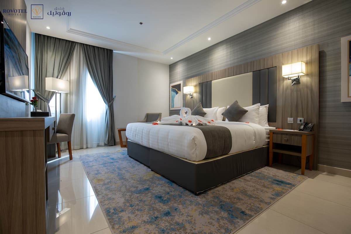 Une chambre d’hôtel moderne avec un grand lit, un bureau et une chaise. On y trouve une lampe de bureau, un téléphone et un luminaire fixé au mur.