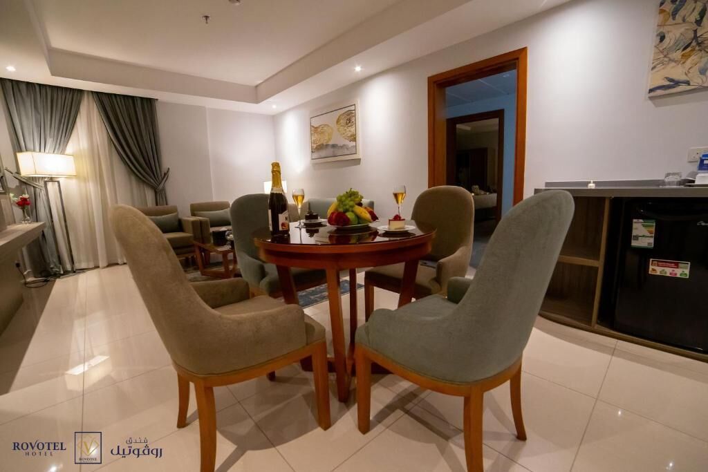 A modern dining area with a round table, four upholstered chairs, a bottle of champagne, and a fruit bowl.