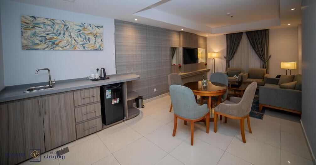A modern hotel room with a kitchen area, a dining table with chairs, a sofa, and a TV mounted on the wall.
