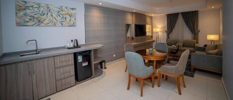 A modern hotel room with a kitchen area, a dining table with chairs, a sofa, and a TV mounted on the wall.