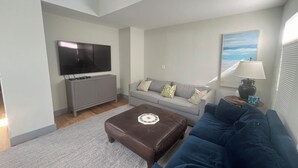 A modern living room with a grey sofa, a brown ottoman, a flat-screen TV mounted on a grey cabinet, and a window with a view of the beach. - Dunes Beach House 1 block from the ocean (Amagansett)
