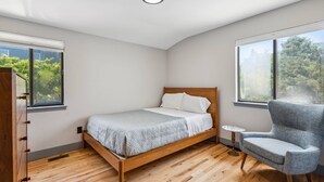 A bedroom with a wooden bed, a grey armchair, and large windows offering a view of trees. - Dunes Beach House 1 block from the ocean (Amagansett)