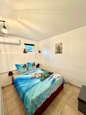 A bedroom with a bed, a window, a wall-mounted air conditioner, a hanging light fixture, and a small decorative item on the wall. - Chez Kelly et Nicolas, le Paradis de Sofaia (Sainte-Rose)
