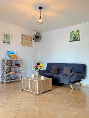 Een woonkamer met een donkerblauwe bank, een houten salontafel met een bloevas, een wandventilator en een plafondlamp.