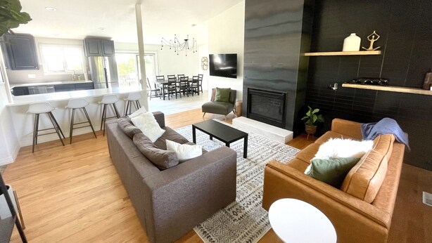 A modern living room with a sofa, coffee table, and a fireplace. There is a kitchen area with a dining table and chairs. - Large Home + Sleeps 20 + Hot Tub + Backyard Oasis + Optional add-on CABOOSE (Denver)