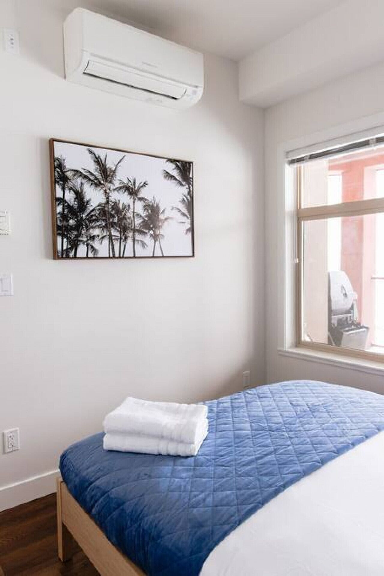 Een slaapkamer met een bed, een wandairconditioner, een lijst met een foto van palmbomen, en een raam met uitzicht op een gebouw.