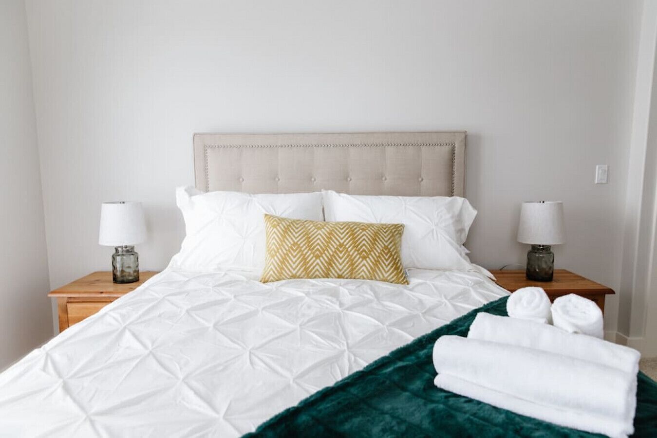 Una cama bien hecha con edredón blanco acolchado, una manta verde y una almohada estampada. Hay dos mesitas de noche con lámparas y toallas dobladas.