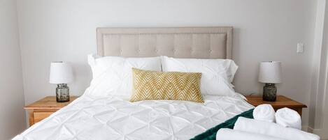 Una cama bien hecha con edredón blanco acolchado, una manta verde y una almohada estampada. Hay dos mesitas de noche con lámparas y toallas dobladas.