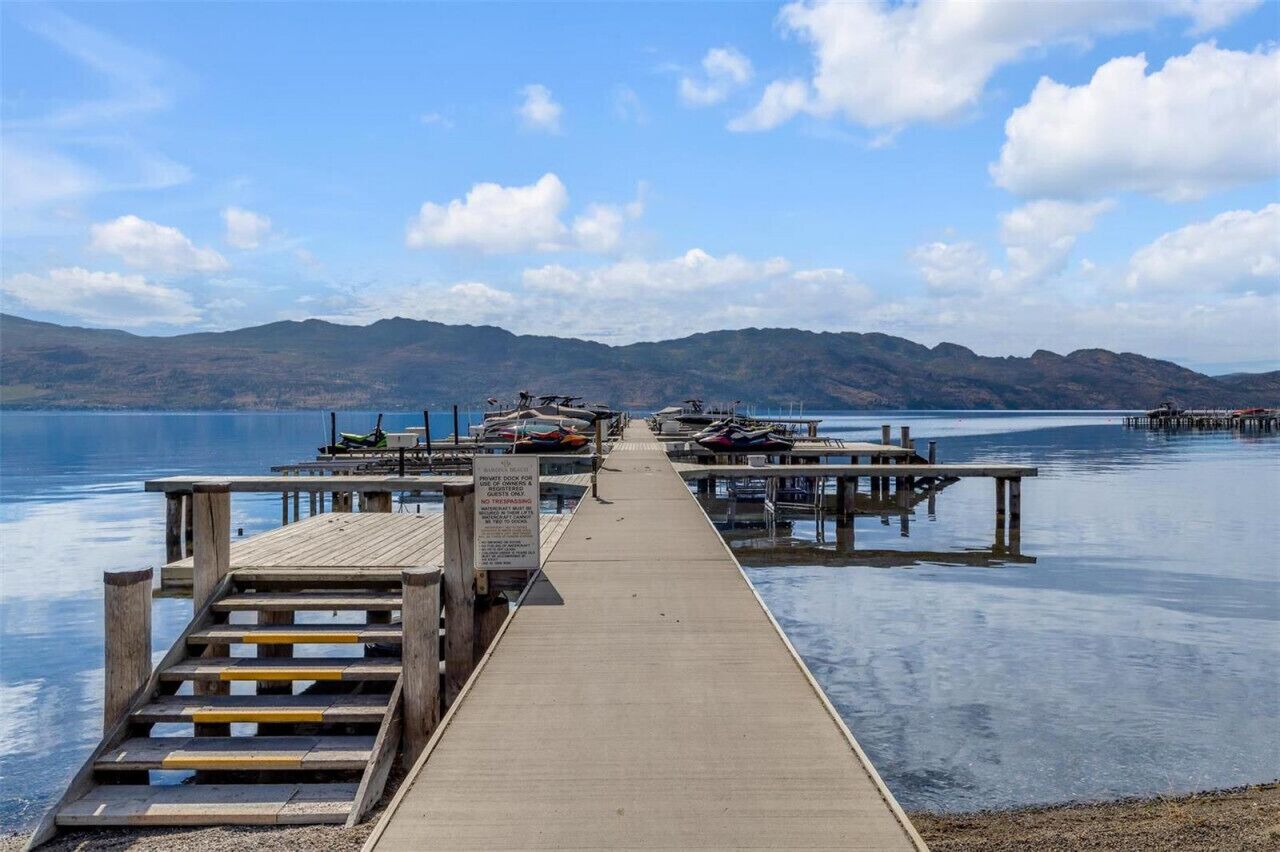 Un muelle de madera que lleva a un embarcadero con barcos, rodeado de aguas tranquilas y montañas al fondo.