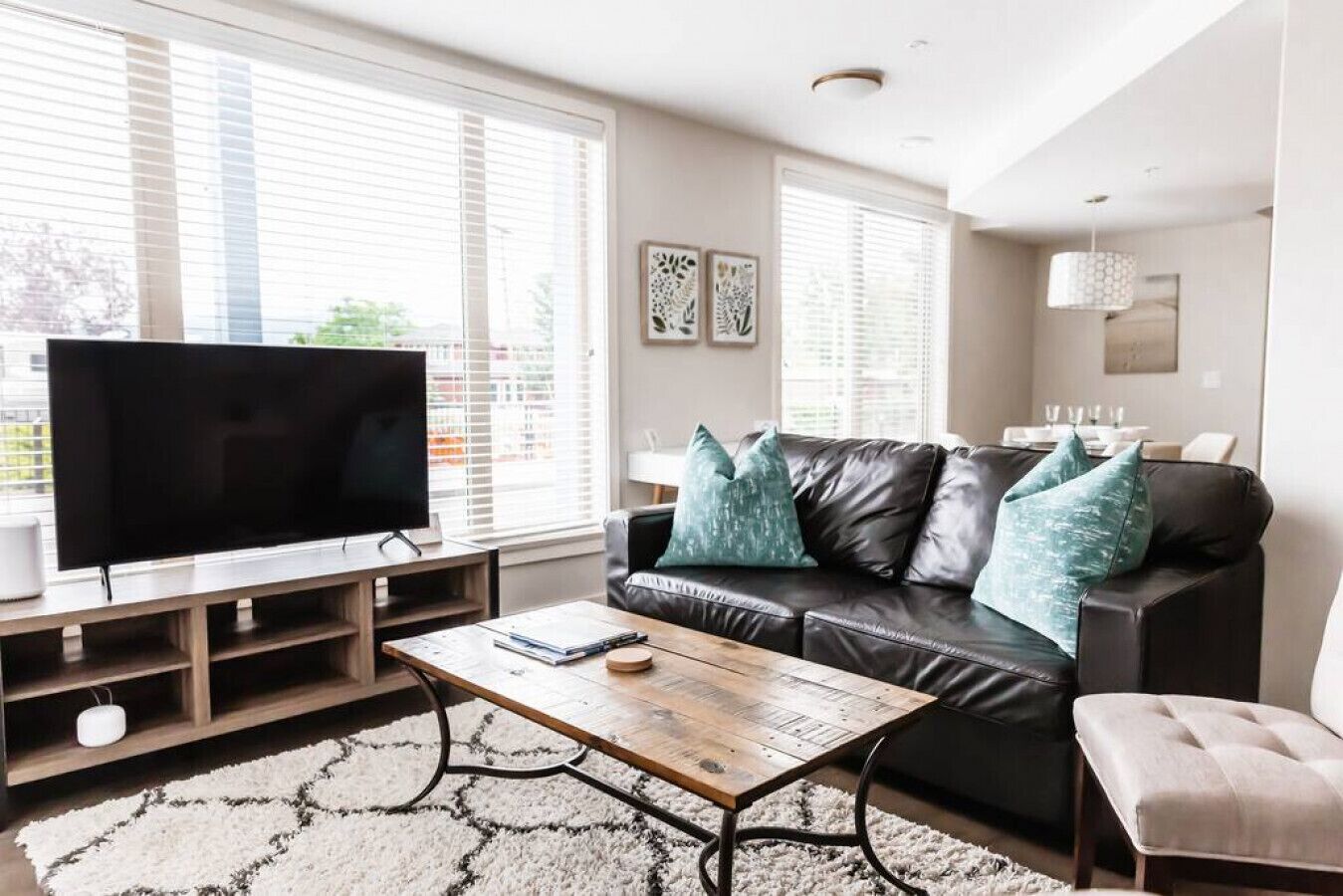 A modern living room with a flat-screen TV, a leather sofa with teal cushions, a wooden coffee table, and a large window with blinds.