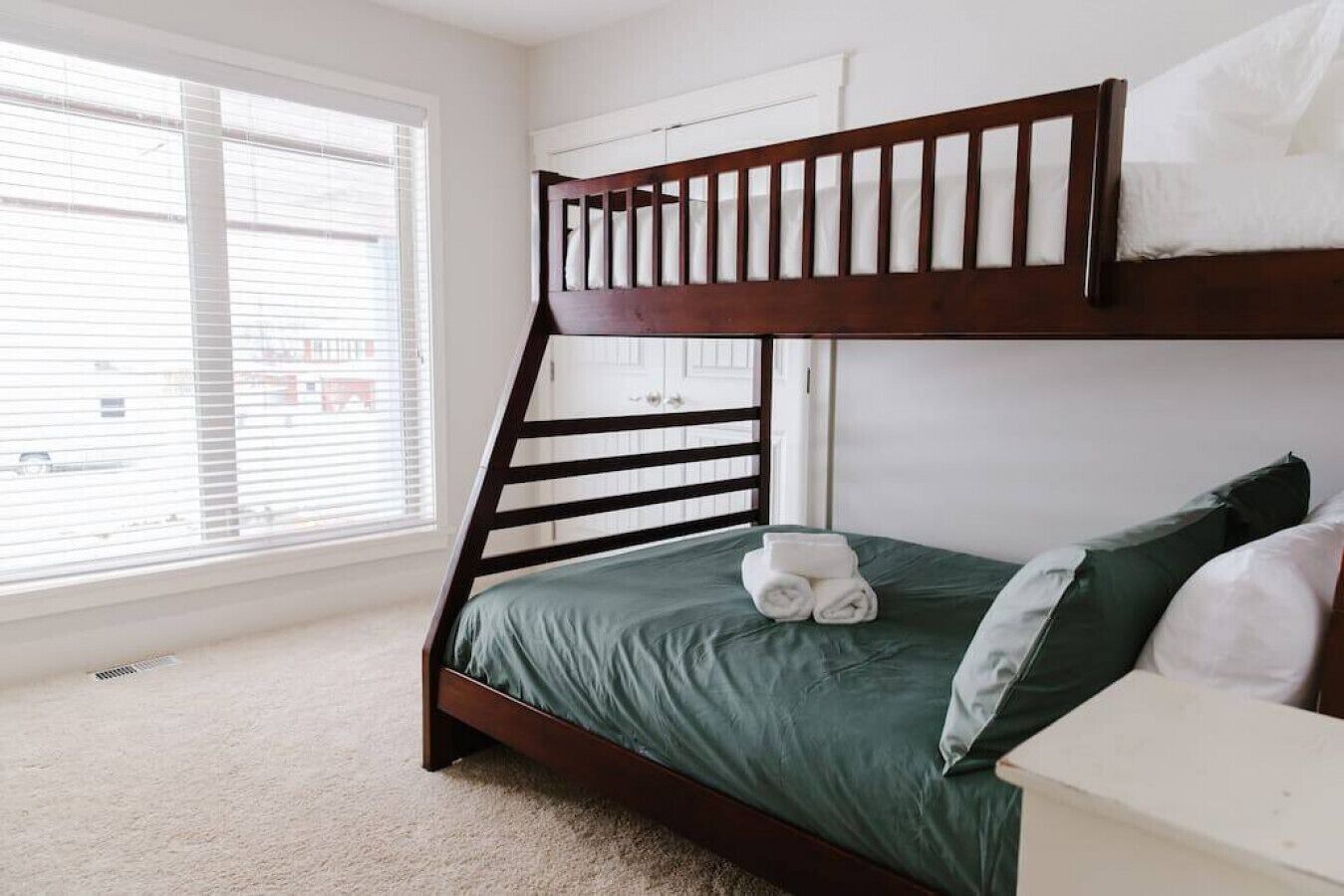 A bunk bed with a green mattress and pillows, a white bedside table, and a window with blinds.