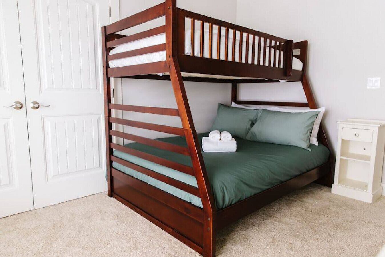 A bunk bed with a wooden frame and green bedding, a white nightstand, and a white door.