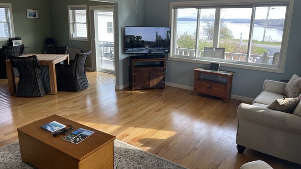 Un salon avec une table basse en bois, un téléviseur, des fauteuils en osier, un canapé et une vue sur un lac, visible à travers de grandes fenêtres.