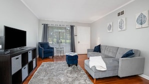 Un salon moderne avec un canapé gris, un fauteuil bleu, un téléviseur à écran plat et une grande fenêtre avec des rideaux.
