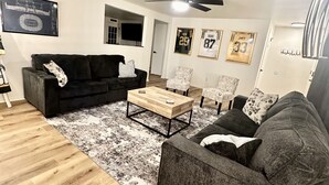 A living room with a black sofa, a wooden coffee table, and a patterned rug. There are framed sports jerseys on the wall and a ceiling fan. - Stay just a short walk -0.3- miles from Lambeau! (Green Bay)