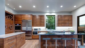Cozinha moderna com armários de madeira, eletrodomésticos de aço inoxidável, uma ilha central com banquetas e uma grande janela que proporciona iluminação natural.
