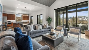 Uma sala de estar ampla com um sofá cinza, uma mesa de centro e vista para uma cozinha e montanhas através de grandes janelas.