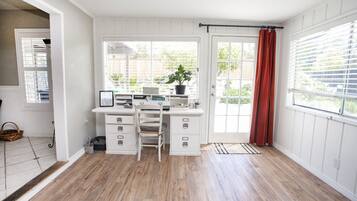 A room with a wooden floor, white walls, a desk with drawers, a chair, a potted plant, and a window with a red curtain.