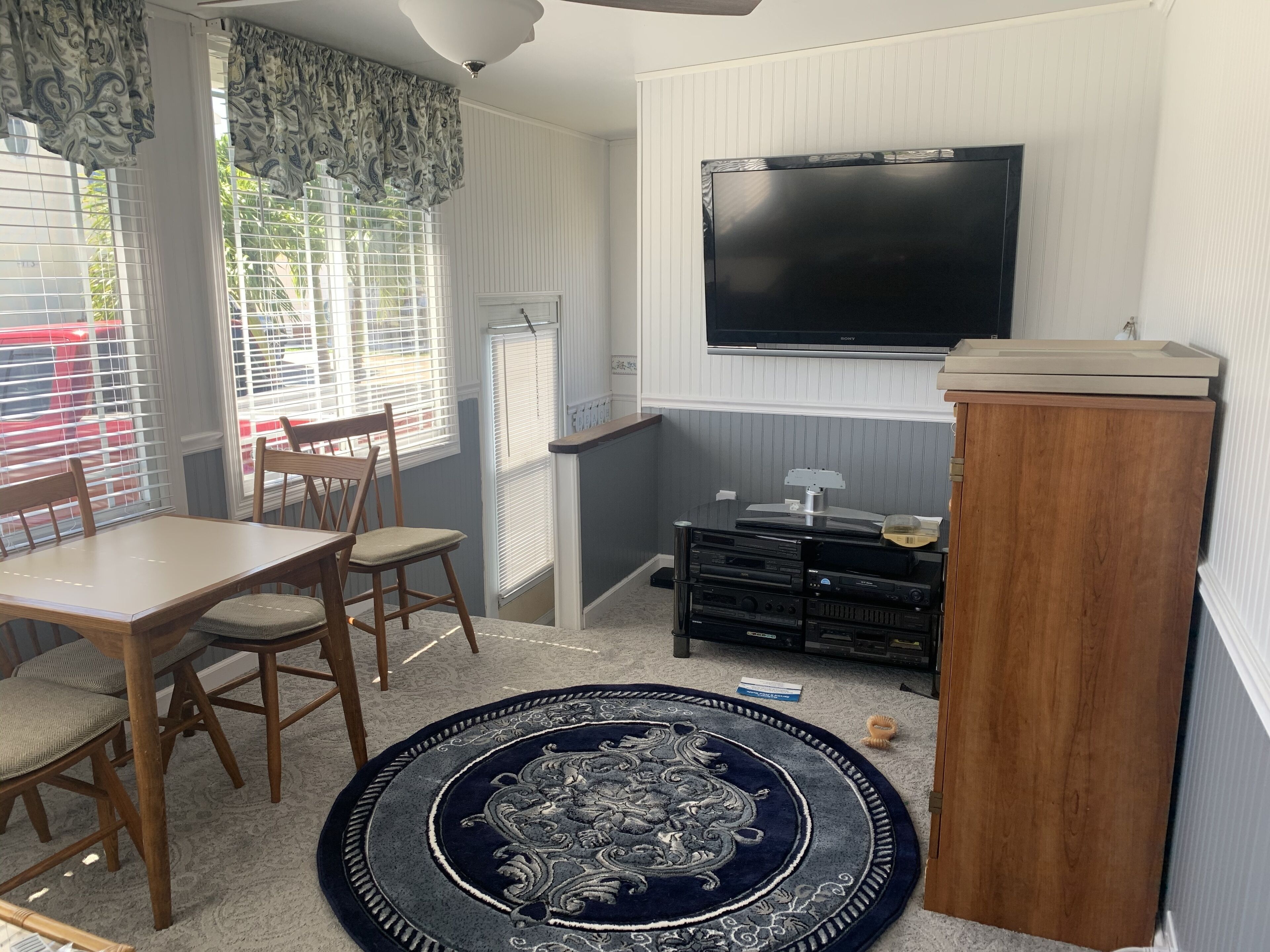 Una habitación con una mesa de comedor, sillas, un televisor montado en la pared, un armario de madera y piso alfombrado.