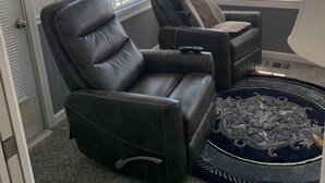 Une pièce d’angle dotée de deux fauteuils inclinables en cuir, d’un tapis à motifs et offrant une vue sur la rue grâce à de grandes fenêtres ornées de lambrequins à motifs.
