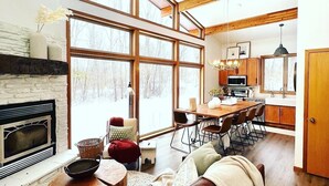 Ein geräumiges Wohnzimmer mit Kamin, großen Fenstern, einer Essecke und einer Küche.