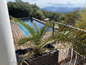 A pool with a wooden deck, surrounded by greenery and mountains in the background.