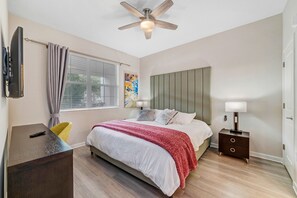 Quarto com cama, mesa de cabeceira, abajur, televisão e ventoinha de teto.