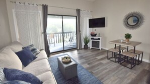 A living room with a sofa, a coffee table, a TV, a dining table with chairs, and a balcony with a railing.