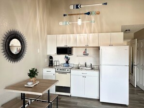 A compact kitchen with white cabinets, a microwave, a stove, and a refrigerator. There is a dining table with benches and a potted plant on the left.