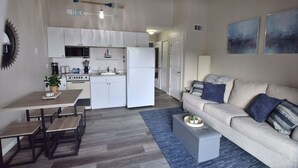 A modern kitchen with white cabinets, a microwave, and a refrigerator. There is a dining table with benches and a small coffee table. The living area has a beige sofa and a blue rug.