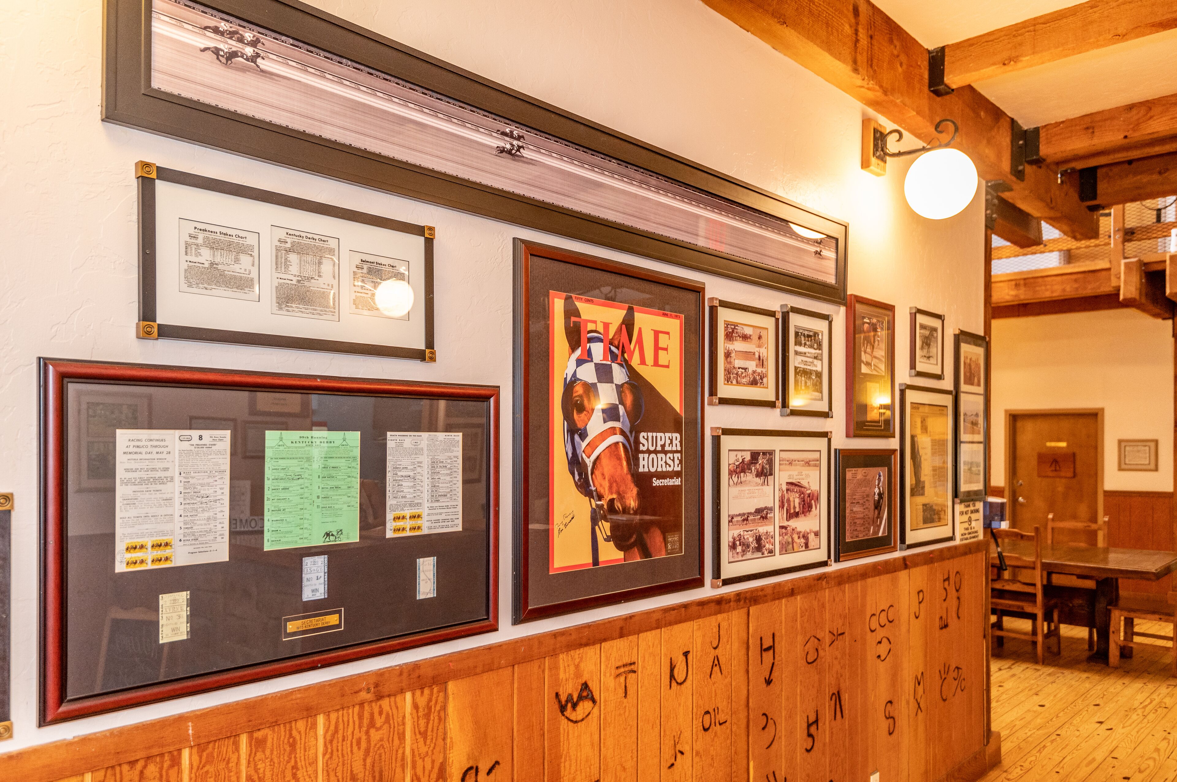 Un mur orné de tableaux et de documents encadrés, un lambris en bois recouvert de graffitis et un plafond en bois avec des poutres apparentes.