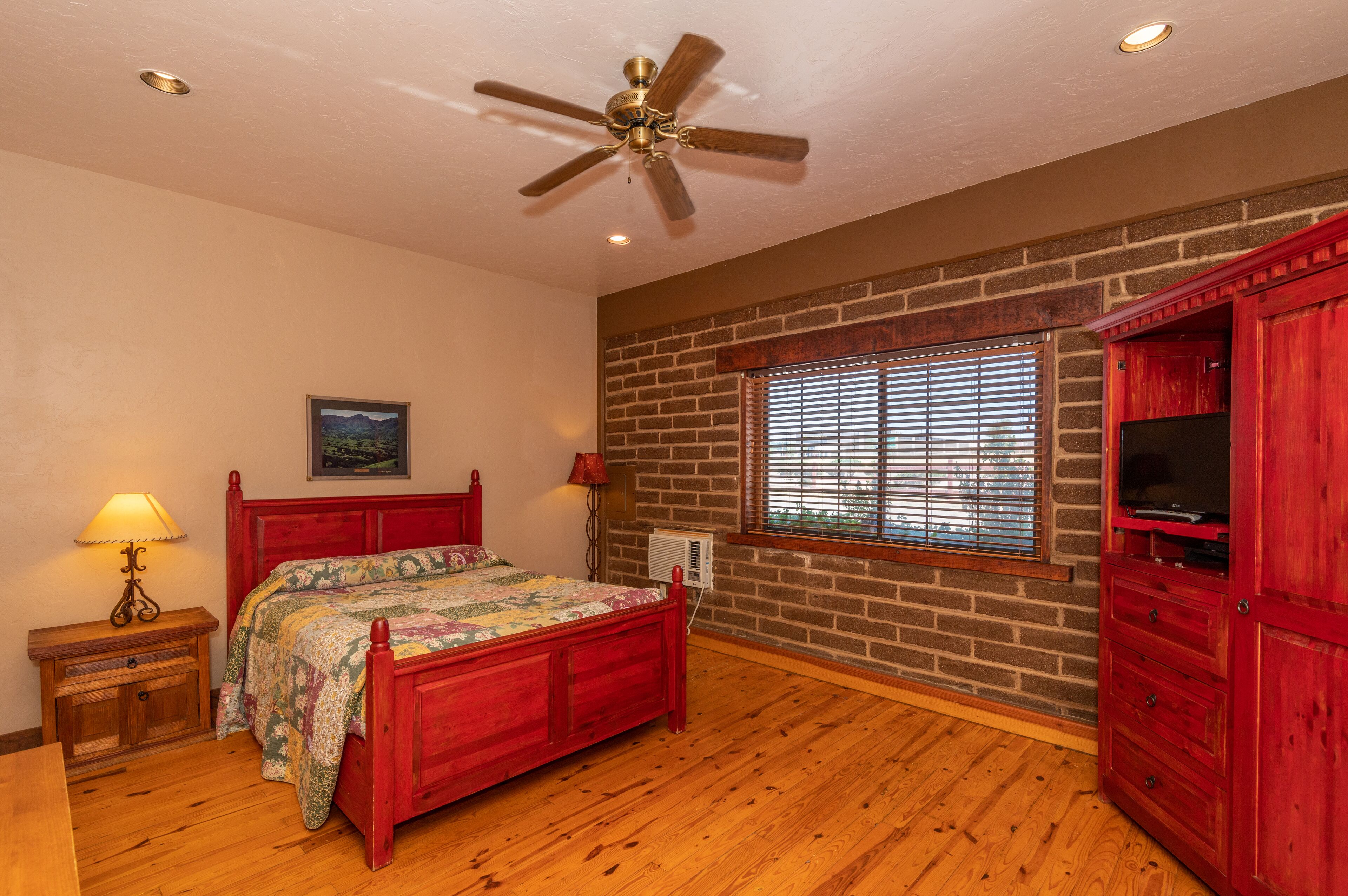 Un dormitorio con piso de madera, una cama roja, ventilador de techo, pared de ladrillo, ventana con persianas y mueble para televisión.