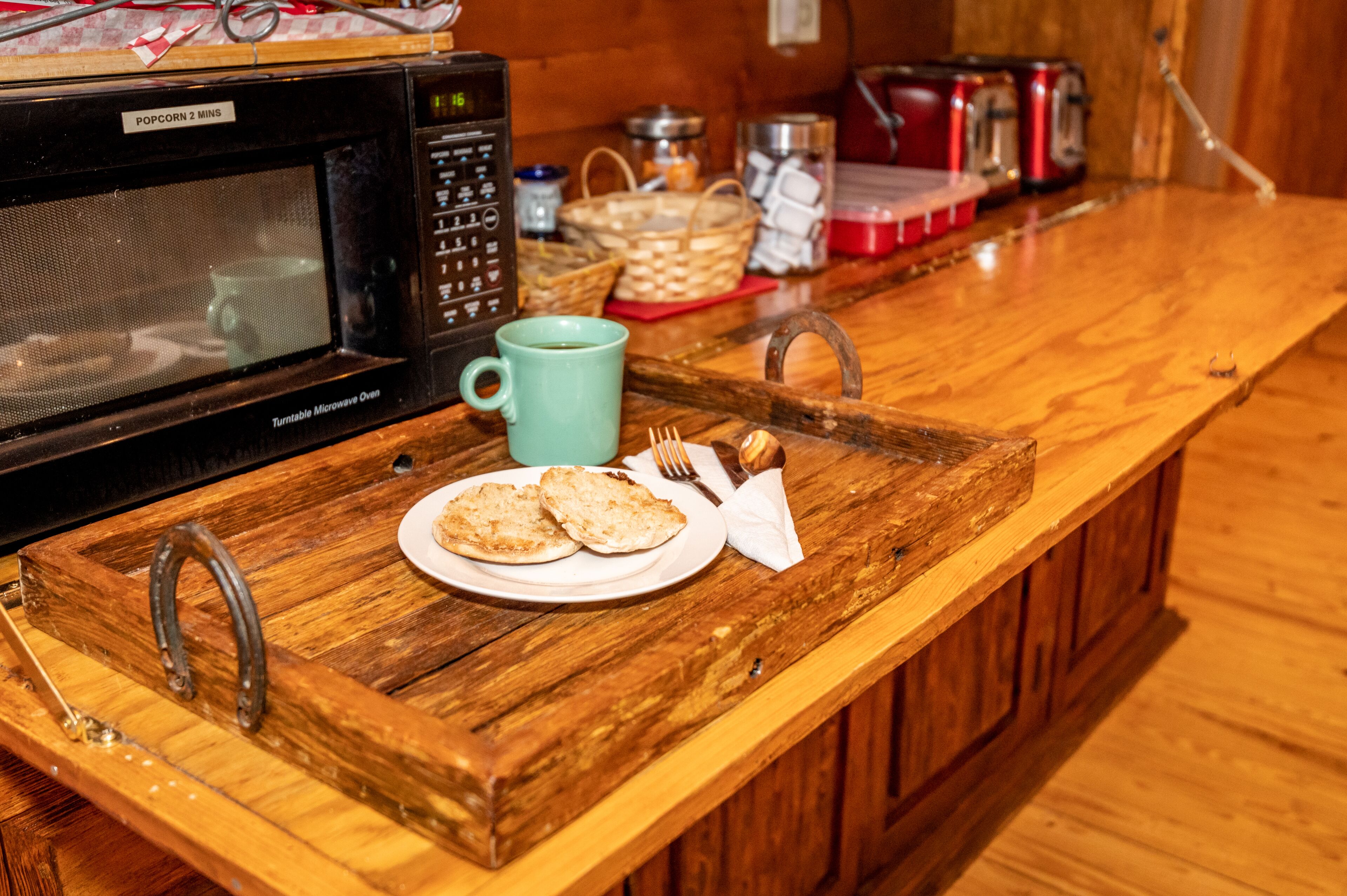 A wooden kitchen counter with a microwave, a tray with bread and a mug, and a basket in the background.