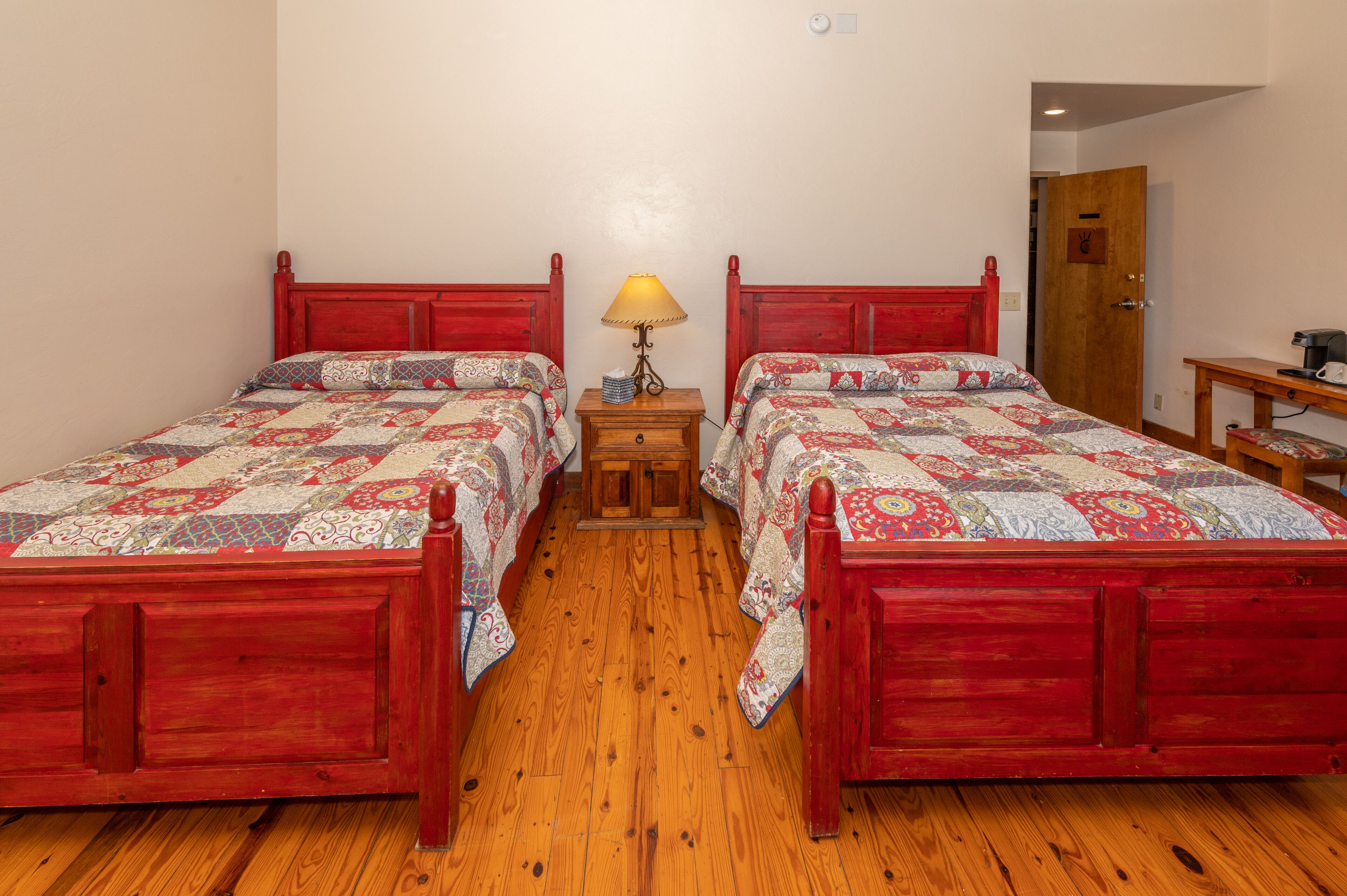A room with two wooden beds with red headboards, a small wooden nightstand with a lamp, and a wooden table with a chair.