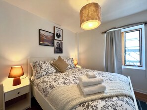A bedroom with a bed, bedside lamps, a hanging light fixture, a window with curtains, and wall art. - Beautiful apartment in the lower town (Québec)