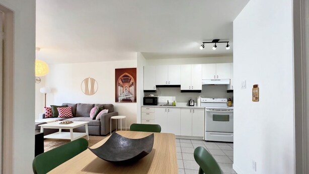 A modern kitchen with white cabinets, a microwave, and an oven. There is a dining area with a wooden table and green chairs. - Beautiful apartment in the lower town (Québec)