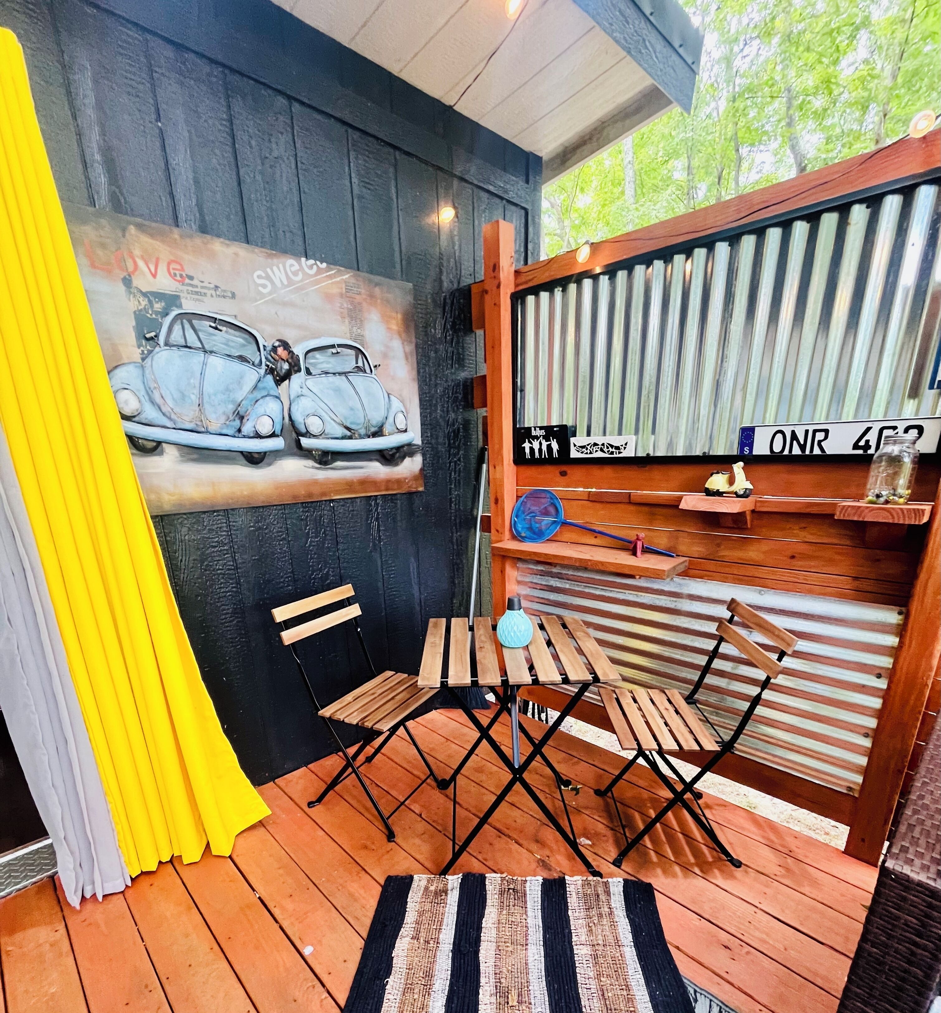En träveranda med en väggmålning som föreställer klassiska bilar, ett litet bord med stolar och en gul gardin.