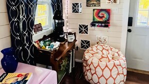 A cozy room with a patterned curtain, a coffee machine, a small table with a book, a bean bag chair, and a wall decorated with various items including a poster and a framed picture.