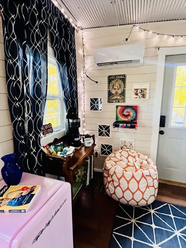 Un cuarto acogedor con una cortina estampada, una cafetera, una mesita con un libro, un sillón informal y una pared decorada con varios objetos, incluyendo un cartel y una fotografía enmarcada.