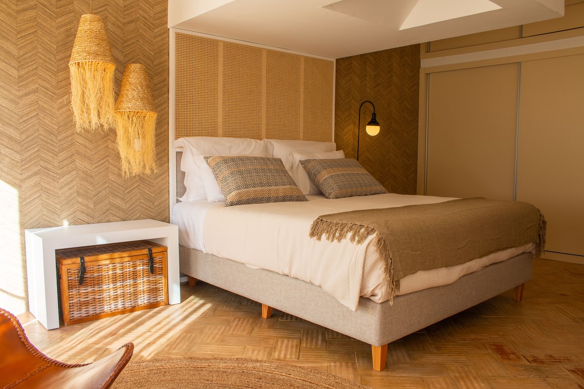 A modern bedroom with a large bed, a bedside table, a woven basket, and a hanging light fixture.