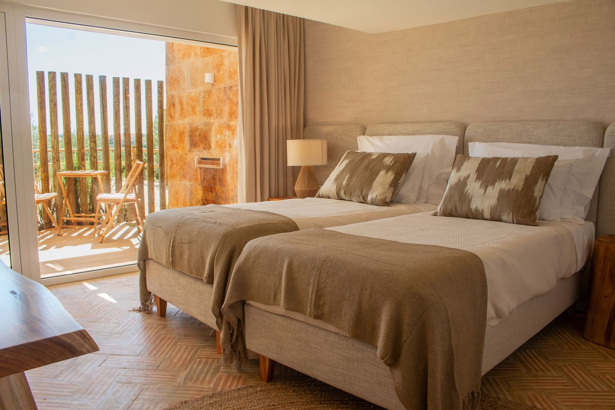 A hotel room with two beds, a wooden floor, a balcony with a view, and a table with chairs.