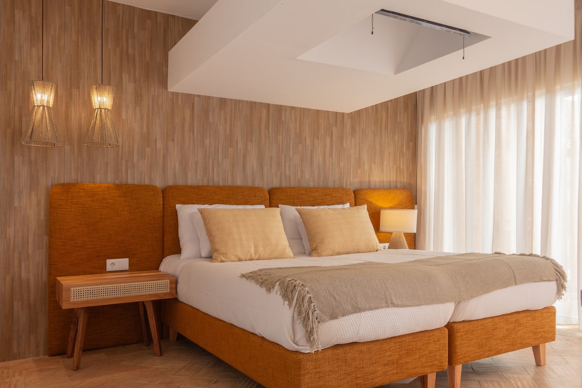 A modern bedroom with a wooden headboard, a large bed with multiple pillows, a bedside table, and a lamp.