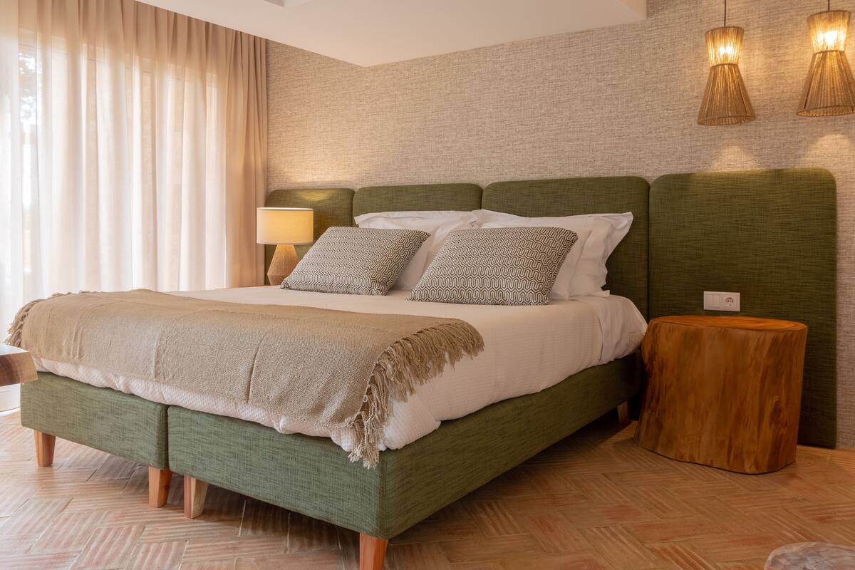 A neatly made bed with a green headboard, a bedside lamp, and a round wooden stool.