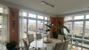 A dining area with a round table and six chairs, large windows with a view of the street, and a potted plant.