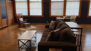 A spacious living room with a dark-colored sofa, a wooden coffee table, and a dining table with chairs. Large windows with blinds are visible, and the room has wooden flooring and a ceiling with a grid pattern. - The Mineral Schoolhouse (Mineral)