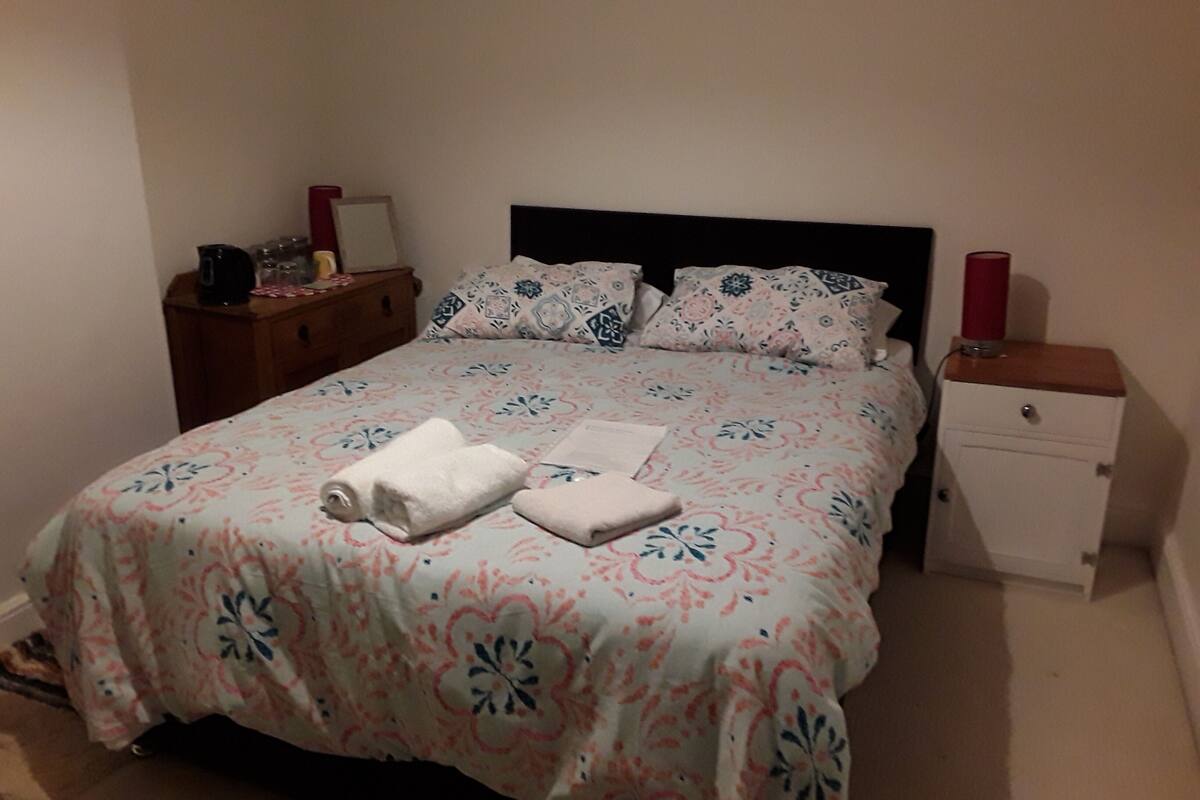 Un lit bien fait, avec une literie à motifs floraux, une table de chevet avec une lampe et une petite table de nuit également équipée d’une lampe.