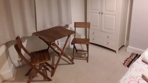 A room with a wooden table and two chairs, a white wardrobe, and a bed with a patterned bedspread.