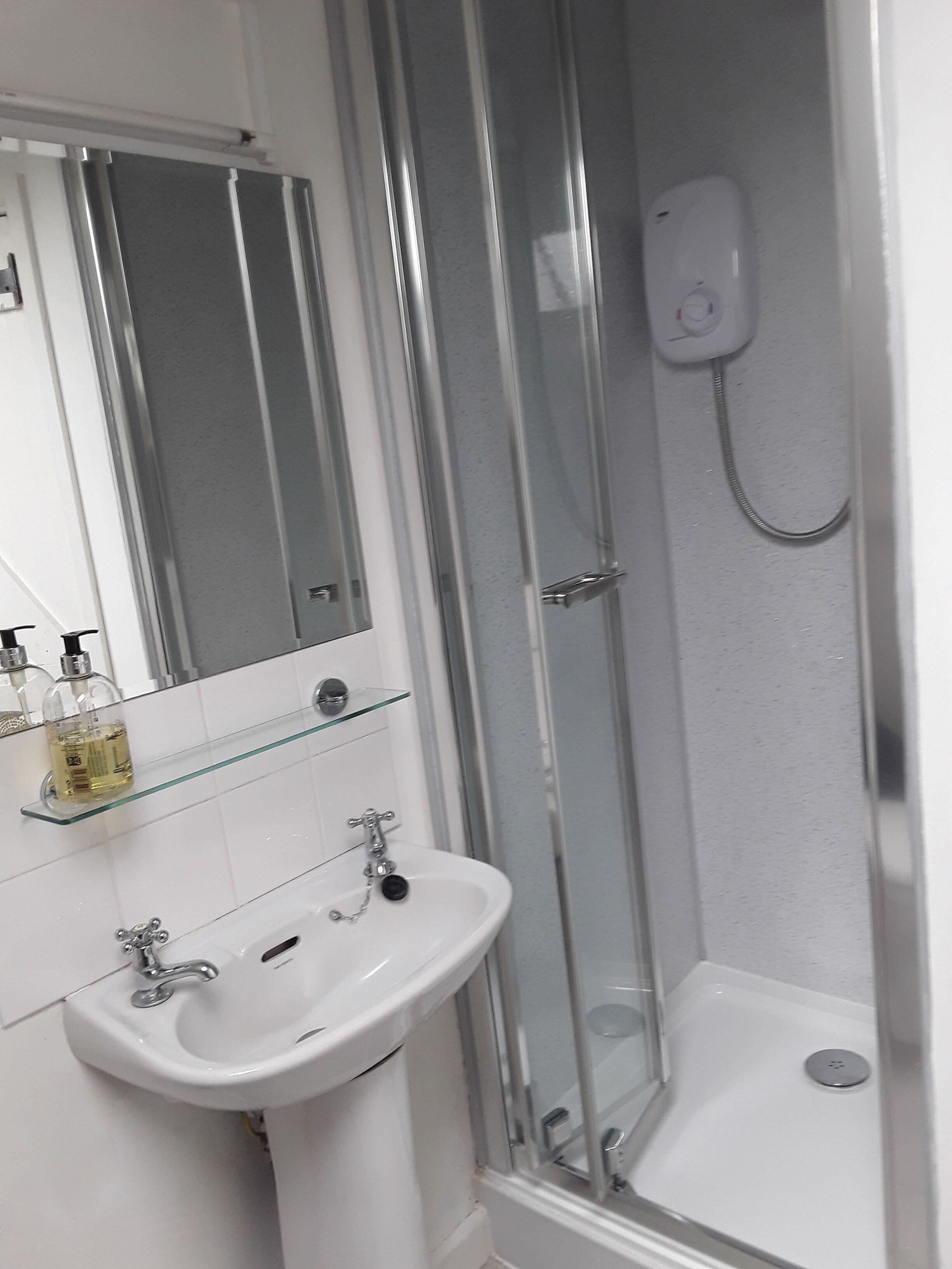 Ein Badezimmer mit Duschkabine aus Glas, einem weißen Waschbecken mit Spiegel darüber und einer Glasablage für Toilettenartikel.