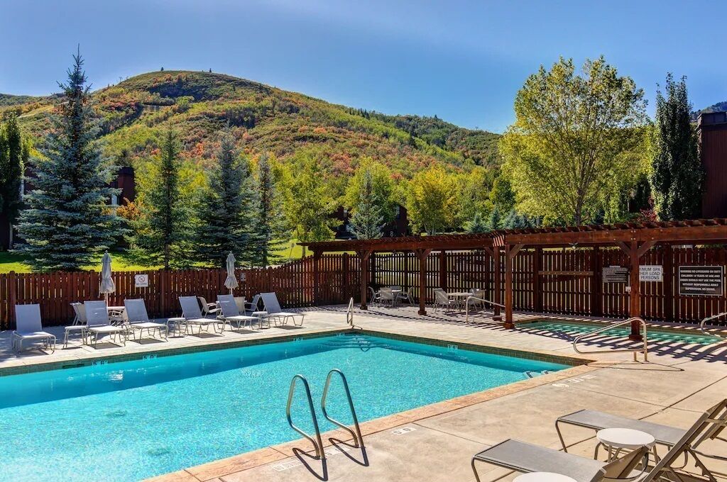 A swimming pool with lounge chairs and a wooden pergola, surrounded by a wooden fence and trees.